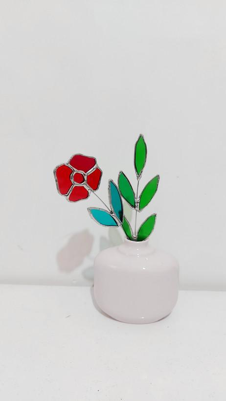 گل شیشه ای قرمز با برگ فیروزه ای و شاخه ای از برگ سبز در گلدان صورتی